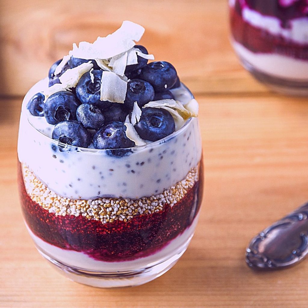 picture of a small glass containing layers of pudding, jam, chia seeds, and blueberries with coconut on a wood table