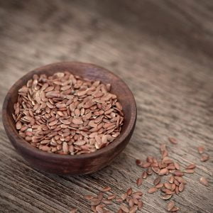 A small wooden bowl of whole flax seeds on lighter wood with flax seeds sprinkled around the bowl